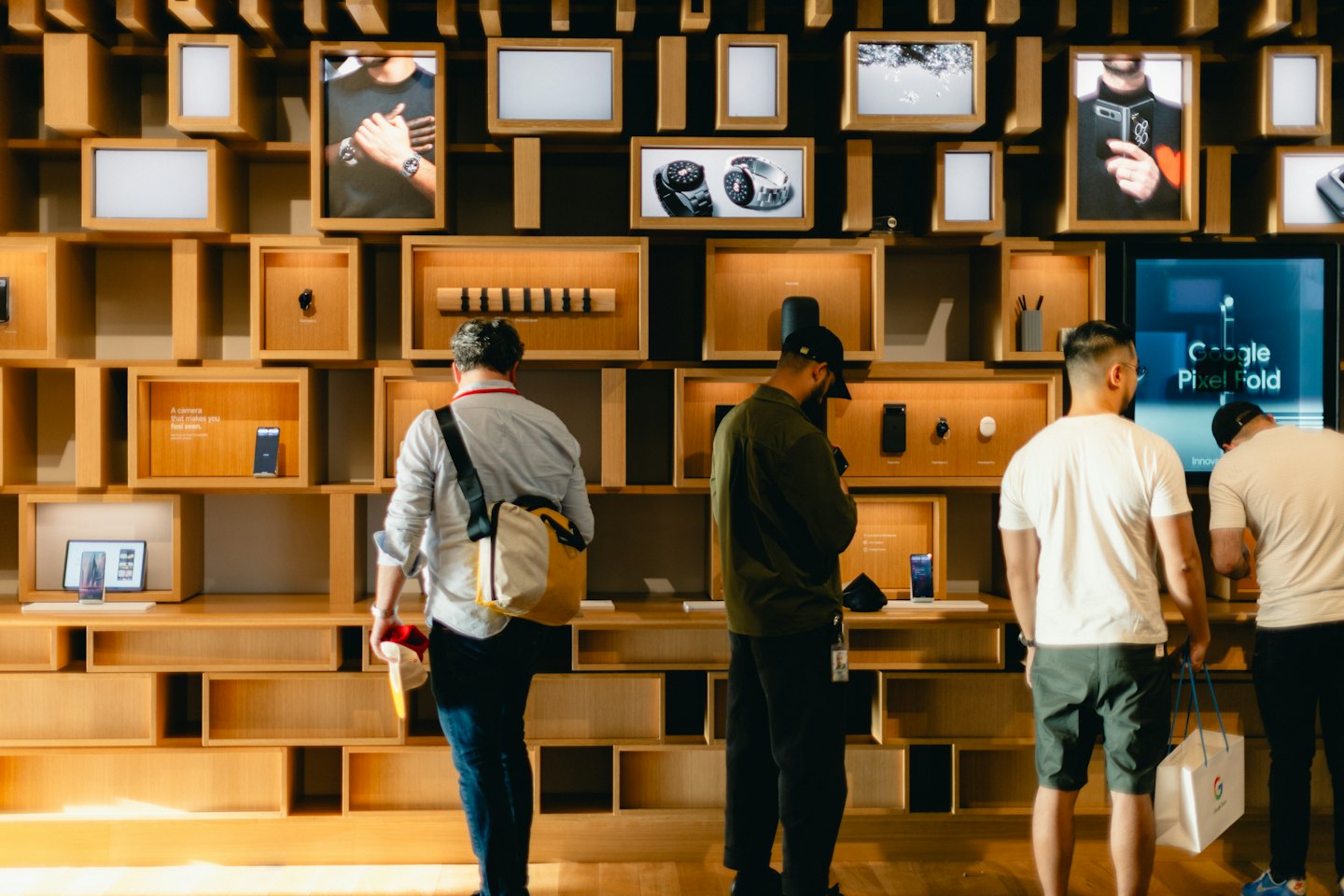 Four men browse electronic devices displayed on a wooden wall.